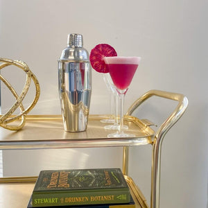 Stainless steel sliver Cobbler Shaker sitting on a gold bar cart with a pink fluffy cocktail in a long stemmed glass garnished next to blood orange garnish