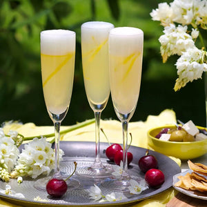 3 flutes of the French 75 cocktail, in dabbled outside light, presented on a tray with fresh cheeries, delicate daffadils a side dish of olives | Cocktail Collective