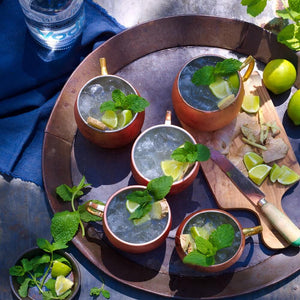Five Moscow Mule cocktails in copper mule mugs with lime and mint garnish on a rustic serving tray outside in the sun