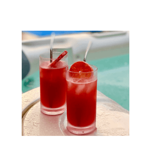2 tall tropical cheery red cocktails in tall glasses garnished with blood oranges & straws, siting beside the pool | Cocktail Collective