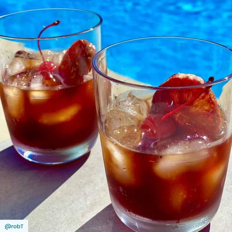 Two Bourbon Old Fashioned cocktails with cherry and  blood orange wheel garnish beside a pool