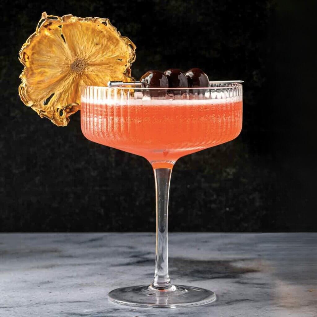 Pink cocktail in a glass with a dried pineapple garnish on a dark background