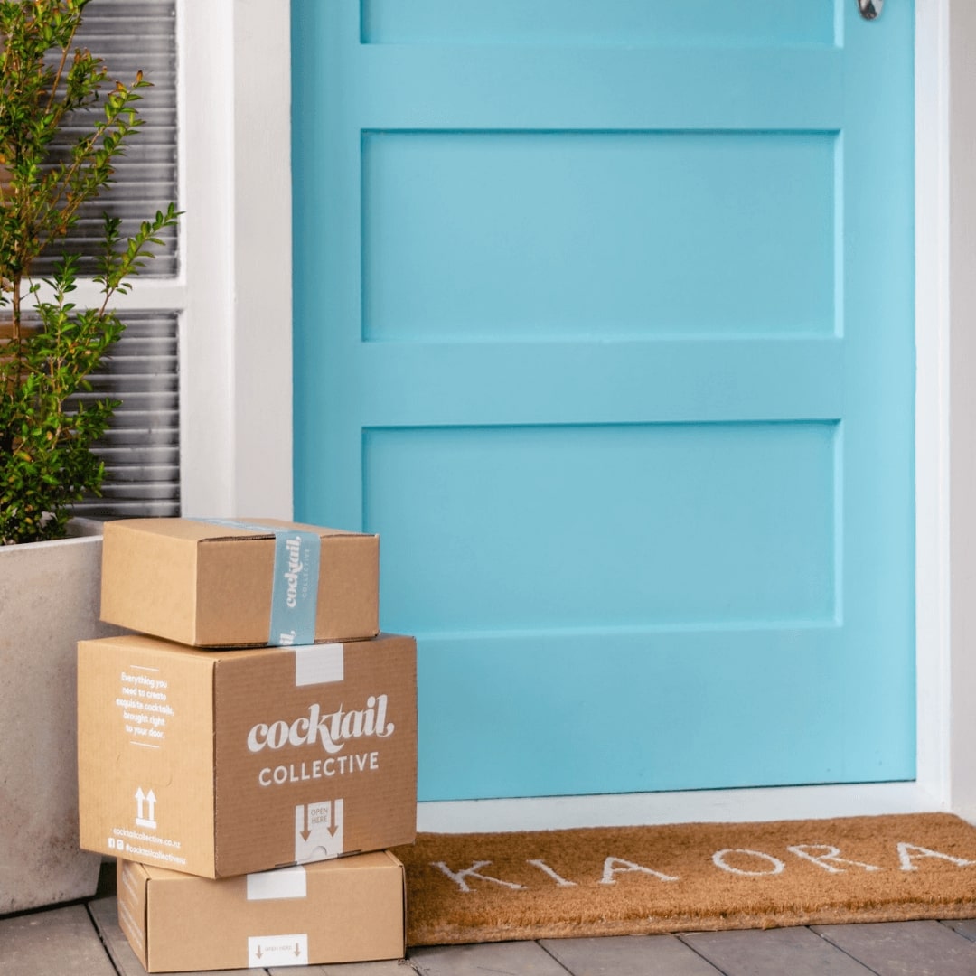 Cocktail Collective gift boxes delivered next to blue door with Kia Ora mat