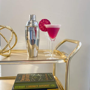 Stainless steel sliver Cobbler Shaker sitting on a gold bar cart with a pink fluffy cocktail in a long stemmed glass garnished next to blood orange garnish 