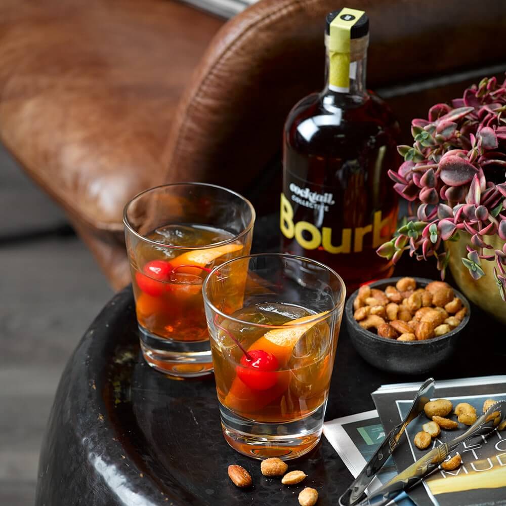 2 Bourbon Old Fashioned cocktails with a bottle of Bourbon on a side table