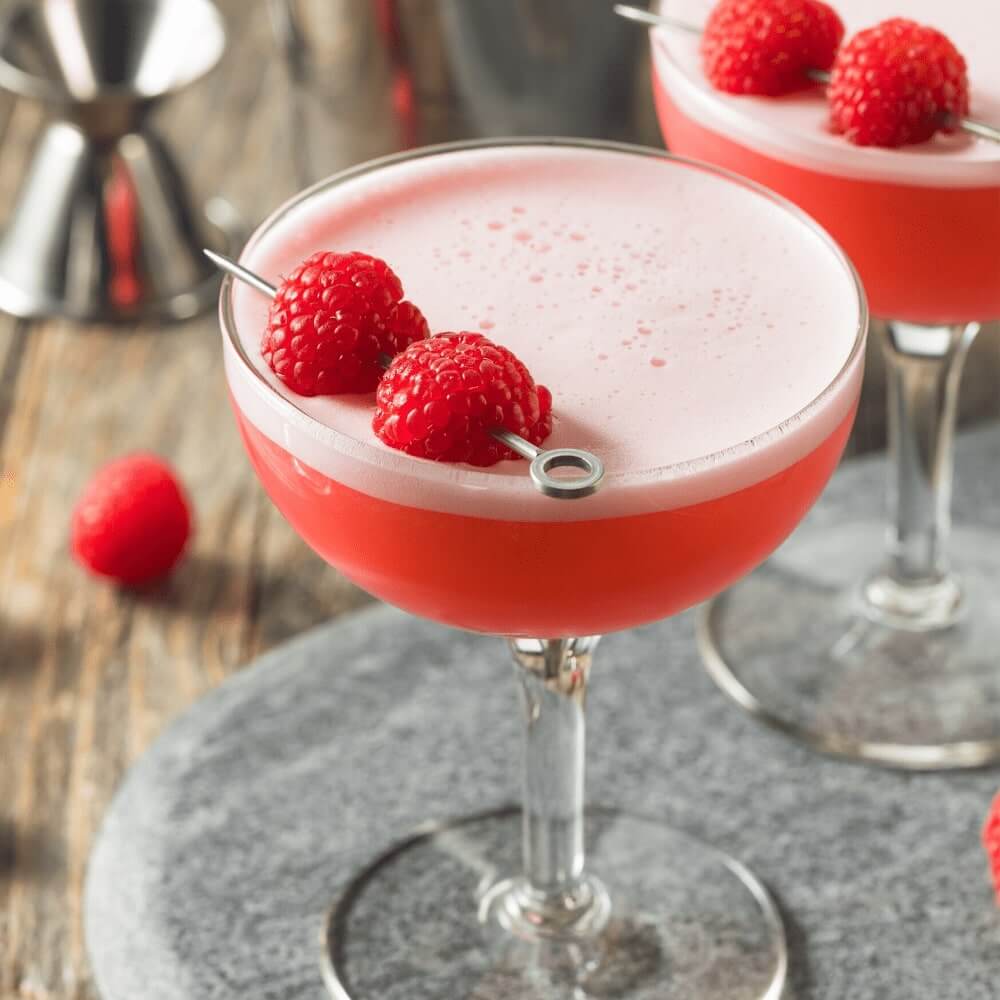 A cocktail coupe filled with the Clover club cocktail & garnished with 2 fresh raspberries on a  silver cocktail stick | Cocktail Collective