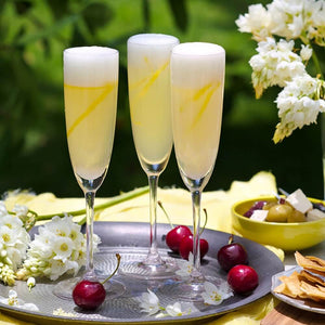 Three champagne flutes of French 75 cocktails sitting on a silver tray with fresh cherries, spring flowers & a bowl of nibbles