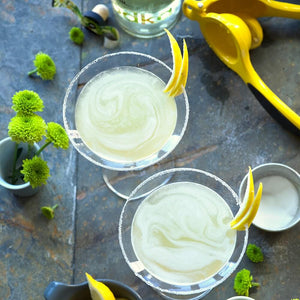 Two Lemon Drop Martini cocktails in sugar-rimmed cocktail coupes, garnished with lemon peels displayed next to a small vase of green flowers