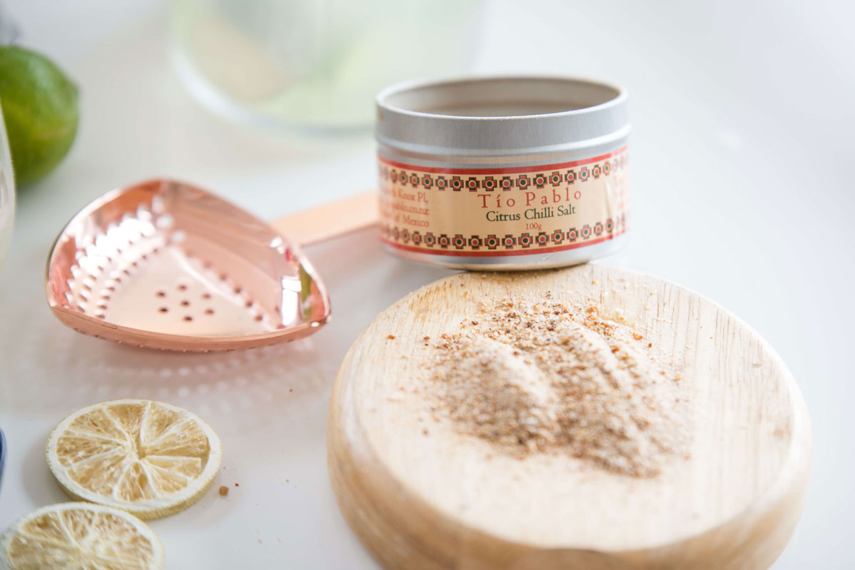 Cocktail garnishes featuring a small open canister of Citrus Chilli Salt from Tio Pablo with loose salt spread on a wooden board next to freeze dried lime wheels from Cocktail Collective
